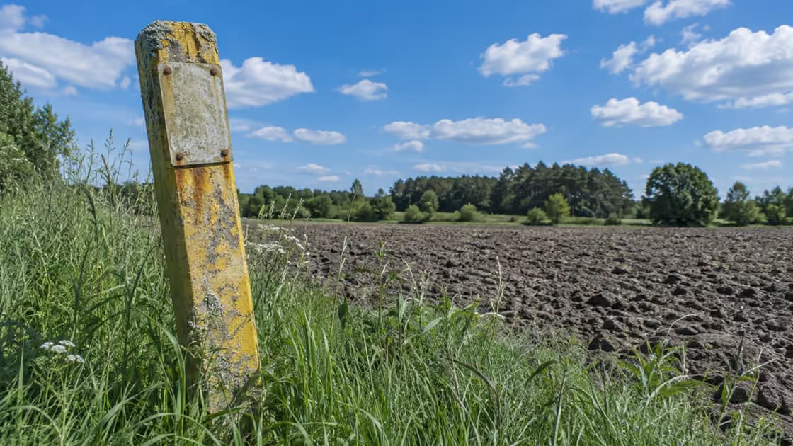 Wysłużony słupek oznacznikowy gazociągu z pustą tabliczką, spękaną żółtą farbą i porostami, w miedzy przy zaoranym polu