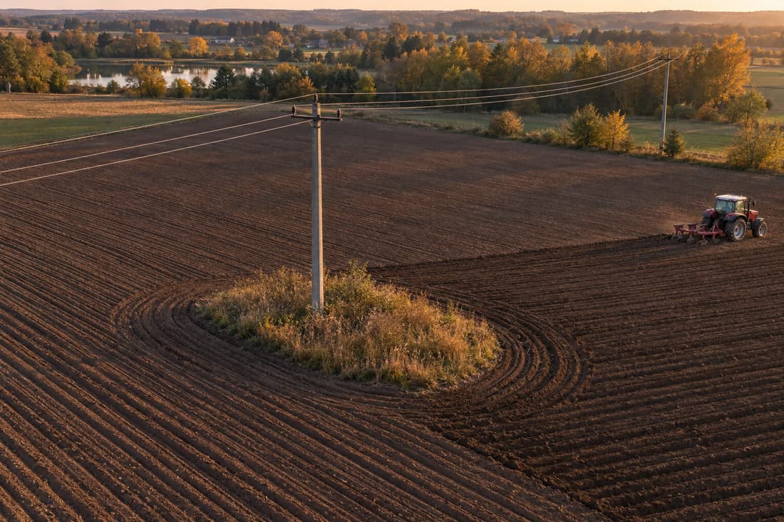 Widok z drona na zaorane pole o zachodzie słońca: bruzdy omijają betonowy słup na środku pola, wokół jego podstawy zostaje kępa suchych chwastów, z prawej ciągnik z pługiem