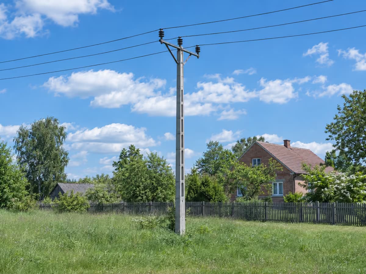Słup średniego napięcia stojący na prywatnej łące, w tle ogrodzenie i zagroda